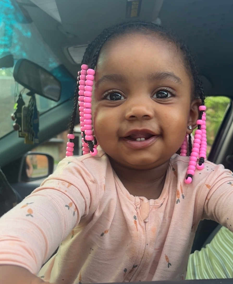 Barbie is registered to the contest to win money with this photo: child, toddler, smiling, braids, pink_beads, face, eyes, clothing, long_sleeves, car_interior, seat, mirror, window, door, person, portrait, cute, happy, young_child, indoors