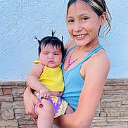 Frida is registered to the contest to win money with this photo: baby, blue_tank_top, child, cute, family, hair, happy, holding, necklace, outdoor, person, pigtails, portrait, skin, smiling, socks, stone_wall, sun_pattern, wall, yellow_clothing