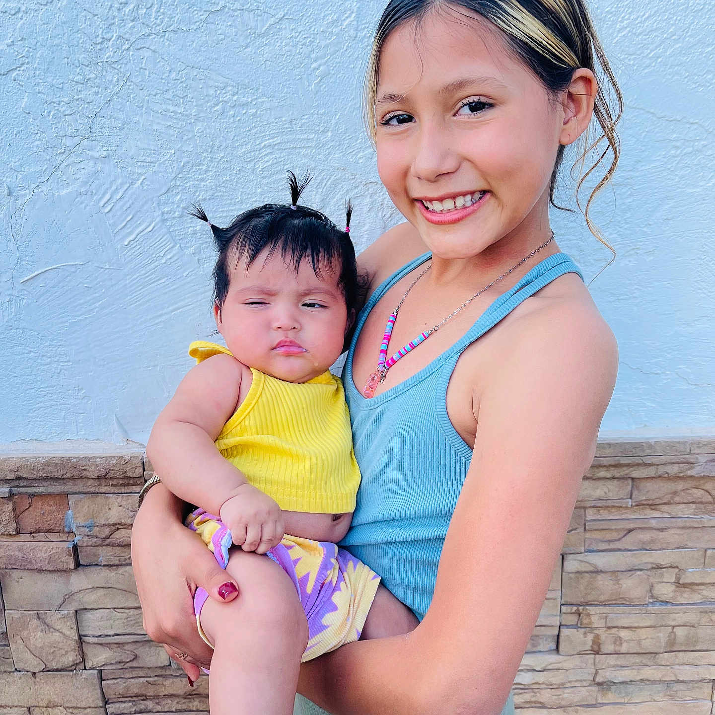 Frida is registered to the contest to win money with this photo: baby, blue_tank_top, child, cute, family, hair, happy, holding, necklace, outdoor, person, pigtails, portrait, skin, smiling, socks, stone_wall, sun_pattern, wall, yellow_clothing
