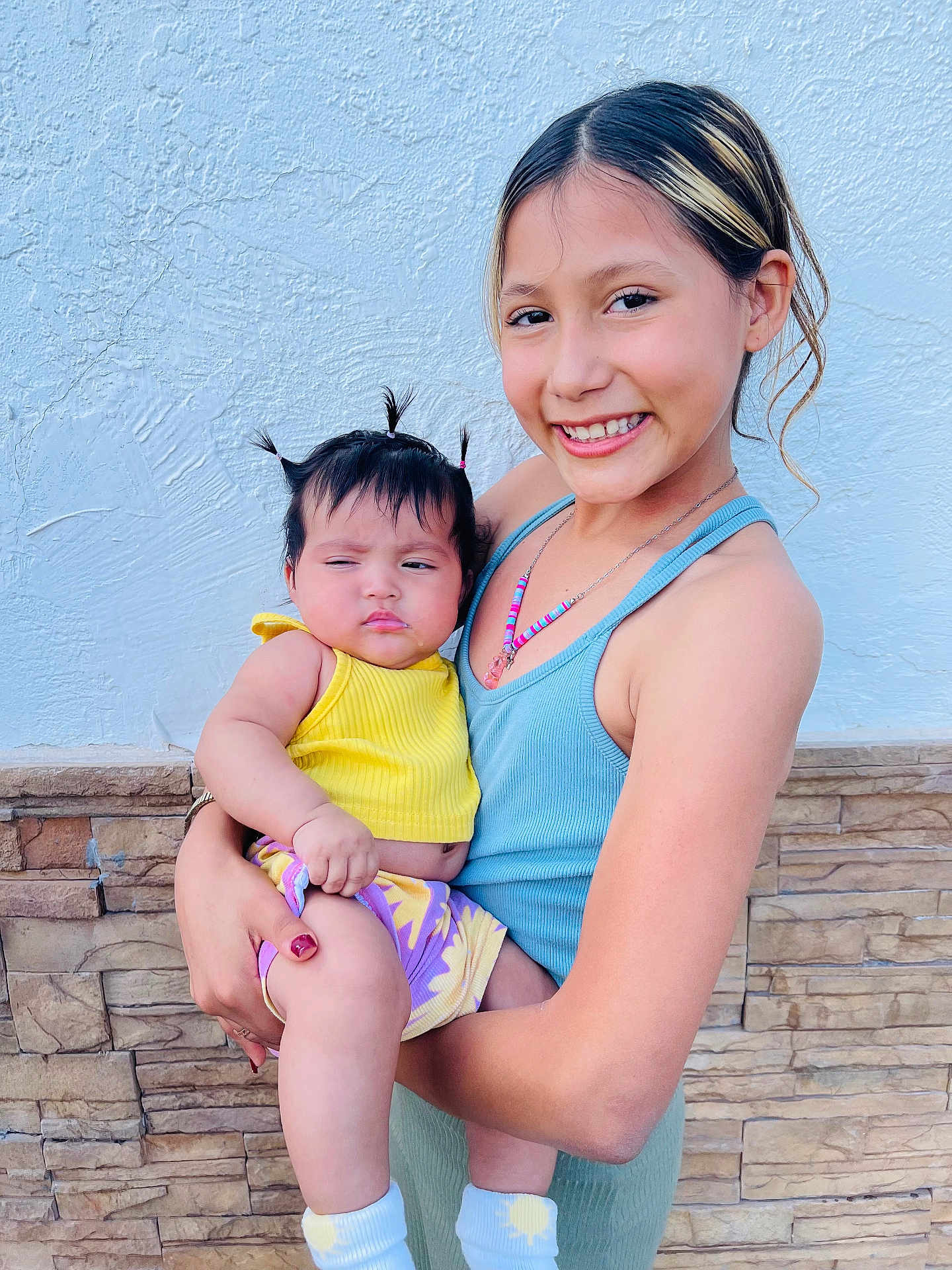 Frida is registered to the contest to win money with this photo: baby, blue_tank_top, child, cute, family, hair, happy, holding, necklace, outdoor, person, pigtails, portrait, skin, smiling, socks, stone_wall, sun_pattern, wall, yellow_clothing