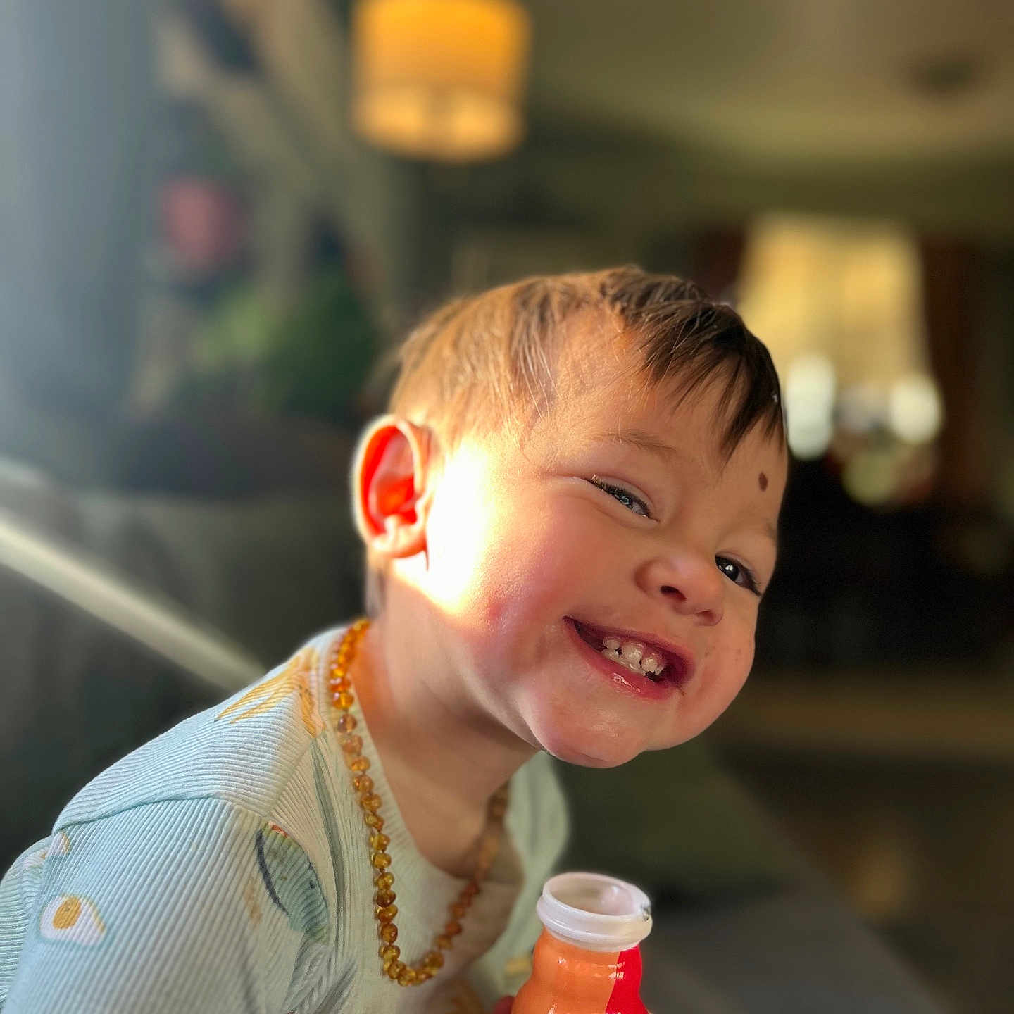 Axyl is registered to the contest to win money with this photo: child, toddler, smiling, happy, face, indoors, bottle, holding, necklace, light, warm_light, background_blur, clothing, portrait, cute, young_child, cheerful, sitting, person, cozy