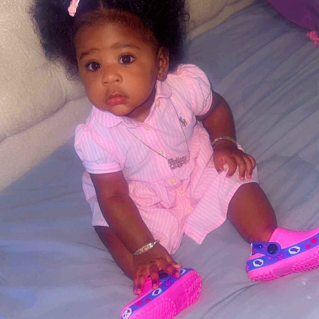 Patience is registered to the contest to win money with this photo: toddler, child, baby, girl, pigtails, pink_bows, pink_dress, colorful_shoes, bed, bedding, headboard, bracelet, necklace, indoor, cute, curious_expression, sitting, person, portrait, soft_lighting
