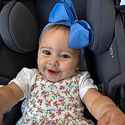 Antonella is registered to the contest to win money with this photo: baby, child, smiling, blue_bow, floral_dress, car_seat, happy, cute, infant, portrait, closeup, young_child, clothing, headwear, person, seat, black_background, reaching_hand, indoors, baby_girl