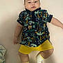 baby, infant, child, lying_down, shirt, shorts, socks, colorful_clothing, patterned_clothing, cute, soft_surface, indoors, young_child, chubby_cheeks, looking_at_camera, hand, leg, footwear, casual_wear, portrait
