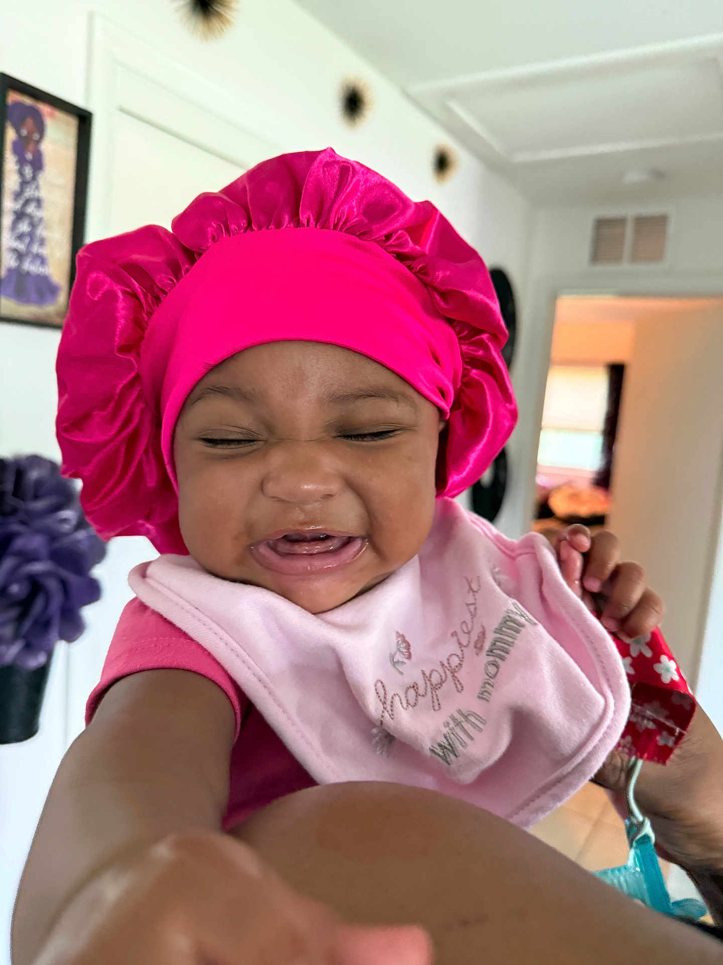 Sareya joined the competition — help win amazing prizes! baby, pink_bonnet, bib, smiling, indoor, child, person, hand, face, clothing, home, portrait, infant, cute, expression, reaching_hand, pink_shirt, wall_art, doorway, decor