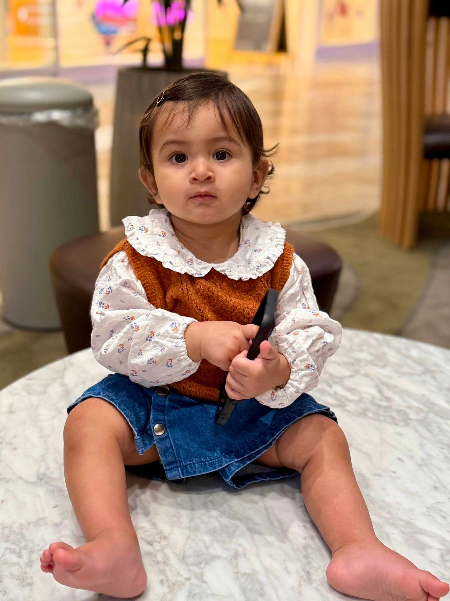 Inayat Kaur joined the competition — help win amazing prizes! toddler, child, indoor, marble_table, remote_control, denim_skirt, vest, long_sleeve_shirt, curly_hair, sitting, barefoot, cute, serious_expression, person, portrait, baby, furniture, floor, blurred_background, curious