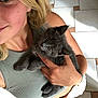 woman, blonde_hair, kitten, gray_cat, indoor, tiled_floor, holding_pet, smiling, casual_clothing, tank_top, shorts, wristband, cute, pet, animal, young_adult, face, hand, closeup, portrait