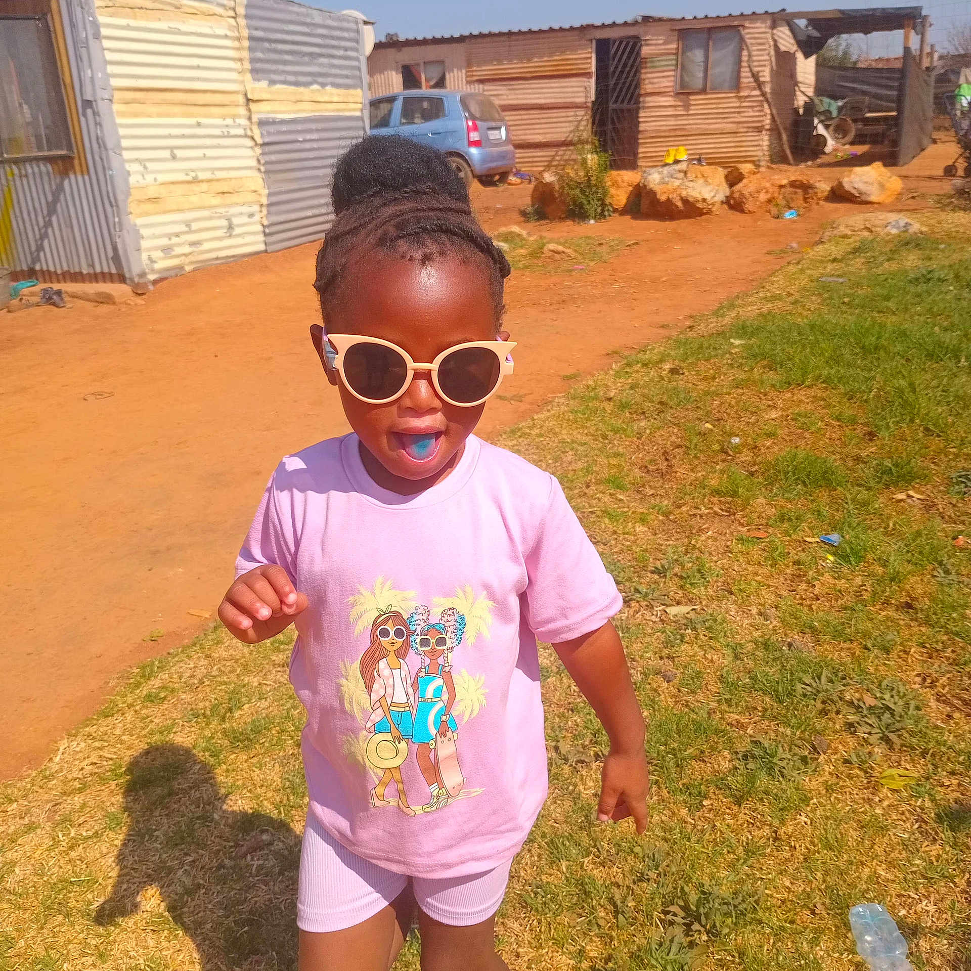 Khanya is registered to the contest to win money with this photo: child, sunglasses, pink_tshirt, outdoor, sunny, grass, dirt_path, house, car, shadow, running, happy, smiling, shorts, footwear, playful, afternoon, hairstyle, casual_clothing, daytime