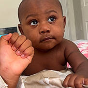 Maurice joined the competition — help win amazing prizes! baby, infant, child, portrait, face, eyes, wide_eyed, sitting, indoors, bedroom, blanket, bedding, adult_hand, hand_holding, brown_skin, bare_chest, short_hair, doorway, home, close_up