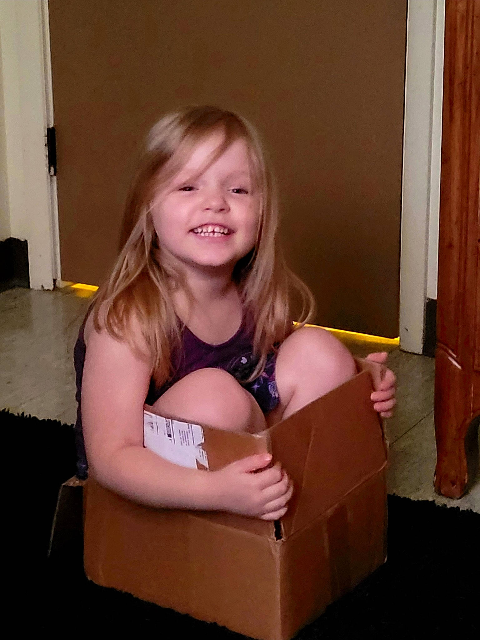 Sylvi is registered to the contest to win money with this photo: blond, box, brown_hair, carton, chest, child, floor, flooring, fun, hairstyle, happy, hardwood, human_leg, joy, packing_materials, person, shipping_box, sitting, smile, thigh