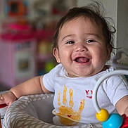 Emma joined the competition — help win amazing prizes! baby, child, infant, smiling, happy, portrait, face, indoors, nursery, activity_seat, toy, colorful, white_shirt, handprint, dark_hair, sitting, playtime, blurred_background, close_up, photograph