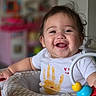 baby, child, infant, smiling, happy, portrait, face, indoors, nursery, activity_seat, toy, colorful, white_shirt, handprint, dark_hair, sitting, playtime, blurred_background, close_up, photograph
