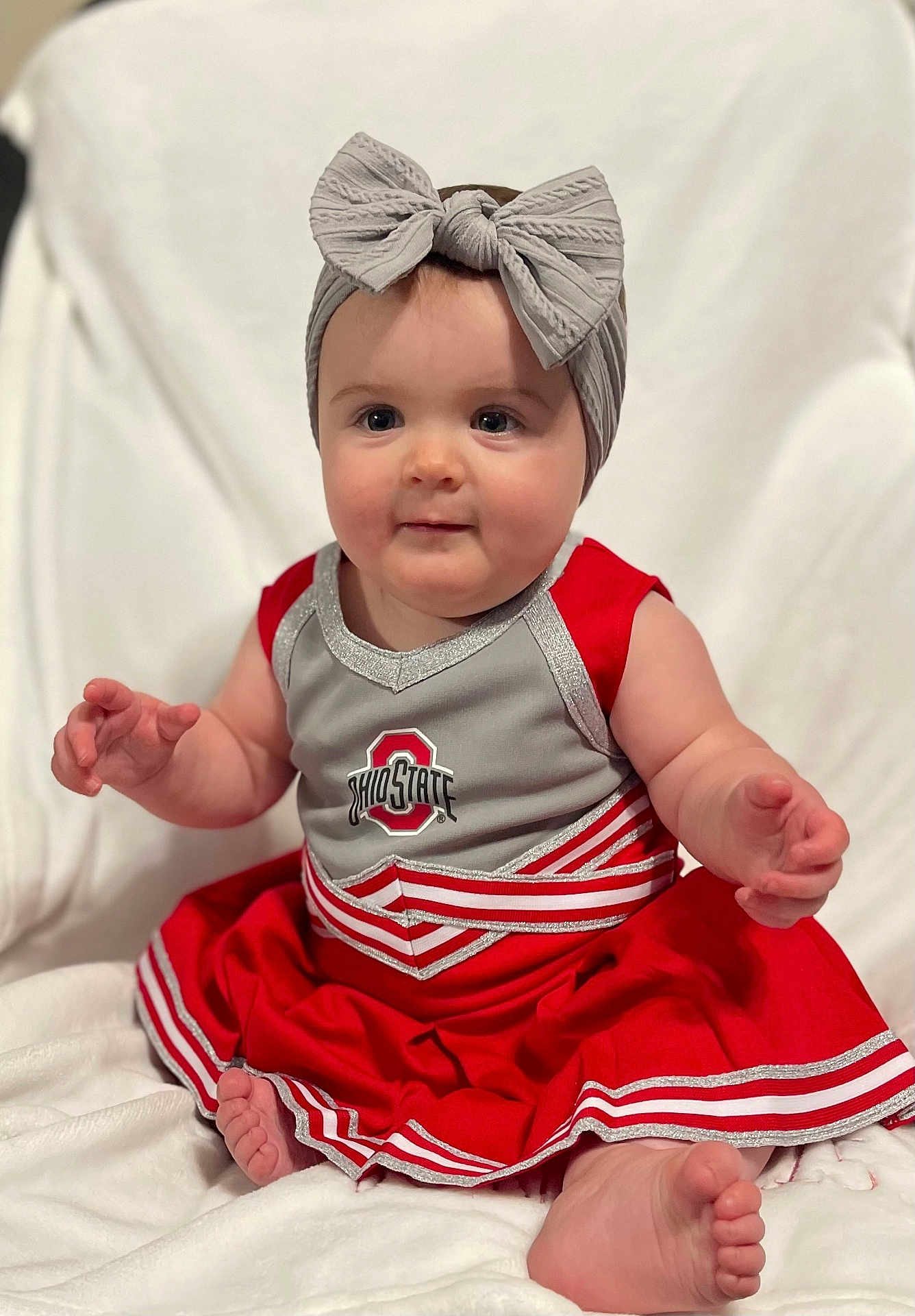Kora is registered to the contest to win money with this photo: baby, child, cheerleader_outfit, headband, bow, red_dress, gray_top, sitting, barefoot, cute, infant, portrait, smiling, indoor, soft_blanket, happy, person, toddler, cheer, sportswear