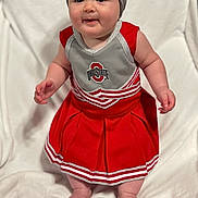 Kora joined the competition — help win amazing prizes! baby, child, cheerleader_outfit, red_dress, headband, bow, blanket, embroidered_text, indoor, cute, smiling, standing, soft_texture, white_background, person, infant, portrait, happy, clothing, footwearless