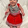 baby, child, cheerleader_outfit, red_dress, headband, bow, blanket, embroidered_text, indoor, cute, smiling, standing, soft_texture, white_background, person, infant, portrait, happy, clothing, footwearless
