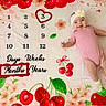 baby, infant, milestone_blanket, pink_clothing, headband, flowers, cherries, smiling, lying_down, months_marker, heart_shape, white_booties, polka_dots, cute, celebration, child, indoors, colorful, portrait, happy