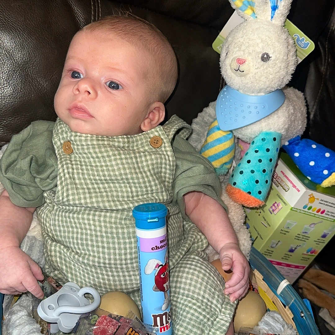 Kolsen joined the competition — help win amazing prizes! baby, child, basket, plush_toy, rabbit, candy, eggs, green_clothing, overalls, leather_couch, indoor, toy, pacifier, sitting, looking_away, soft_toy, colorful, cute, infant, portrait