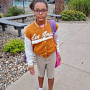 Ivy is registered to the contest to win money with this photo: child, outdoor, person, glasses, jacket, shorts, socks, sandals, bench, plants, sidewalk, fence, pool, backpack, casual_clothing, daytime, standing, young, curly_hair, confident