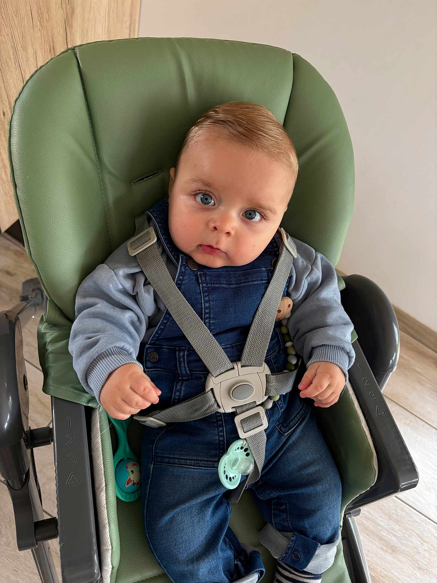 Sandro participe au concours pour gagner de l'argent avec cette photo : baby, child, high_chair, denim_overalls, pacifier, toy, blue_eyes, grey_sweater, straps, indoor, floor, wooden_floor, curious_expression, seated, infant, cute, clothing, person, face, hand