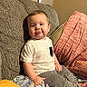 baby, blanket, brown_hair, child, couch, cozy, face, furniture, gray_pants, home, indoor, infant, person, photography, pillow, portrait, sitting, smiling, sofa, white_shirt