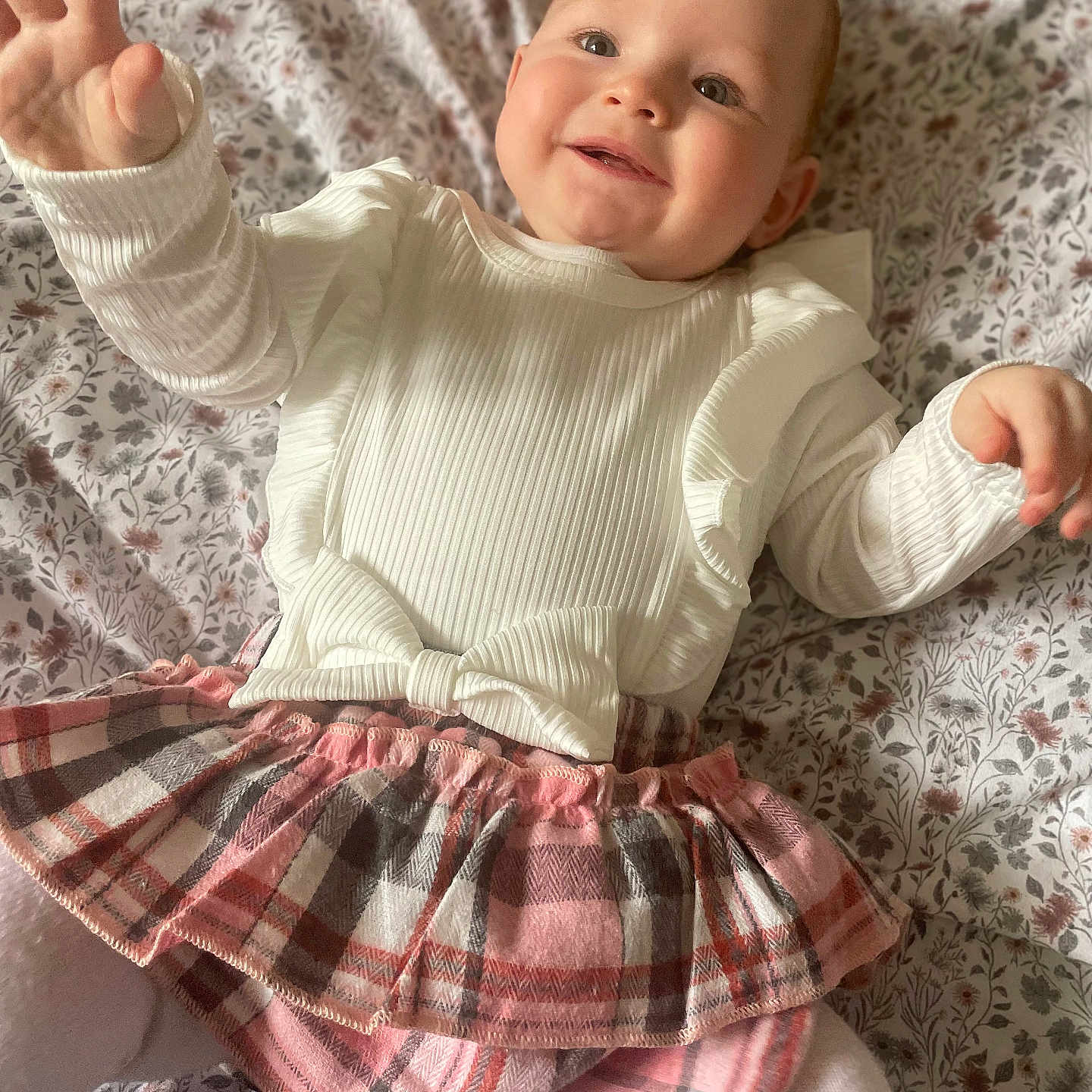 River-Lavinia is registered to the contest to win money with this photo: baby, bedspread, child, cozy, cute, floral_pattern, happy, indoors, infant, lying_down, person, plaid_skirt, portrait, reaching_hand, ruffles, smiling, soft_lighting, toddler, white_clothing, young_child