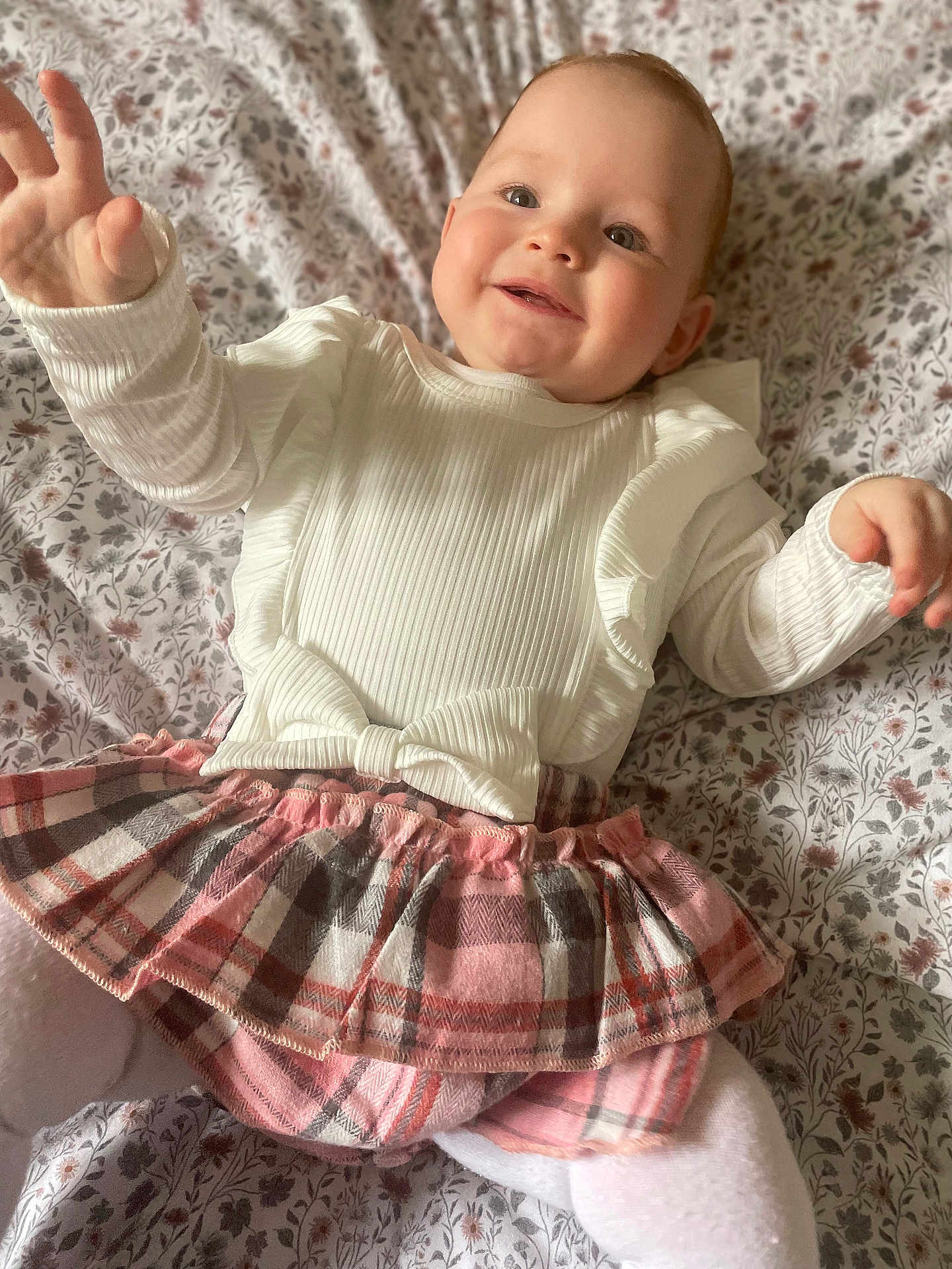 River-Lavinia is registered to the contest to win money with this photo: baby, child, smiling, white_clothing, plaid_skirt, ruffles, lying_down, bedspread, floral_pattern, cute, happy, reaching_hand, infant, soft_lighting, indoors, person, portrait, young_child, toddler, cozy
