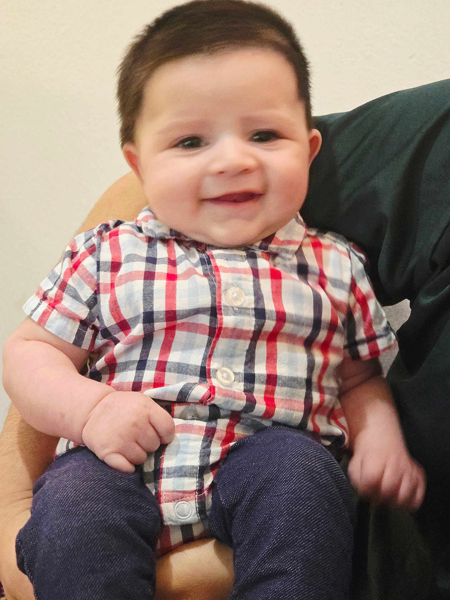Mohammad is registered to the contest to win money with this photo: baby, child, smiling, plaid_shirt, pants, person, indoor, holding, happy, cute, portrait, infant, face, skin, hand, sitting, clothing, adult_arm, background_wall, casual