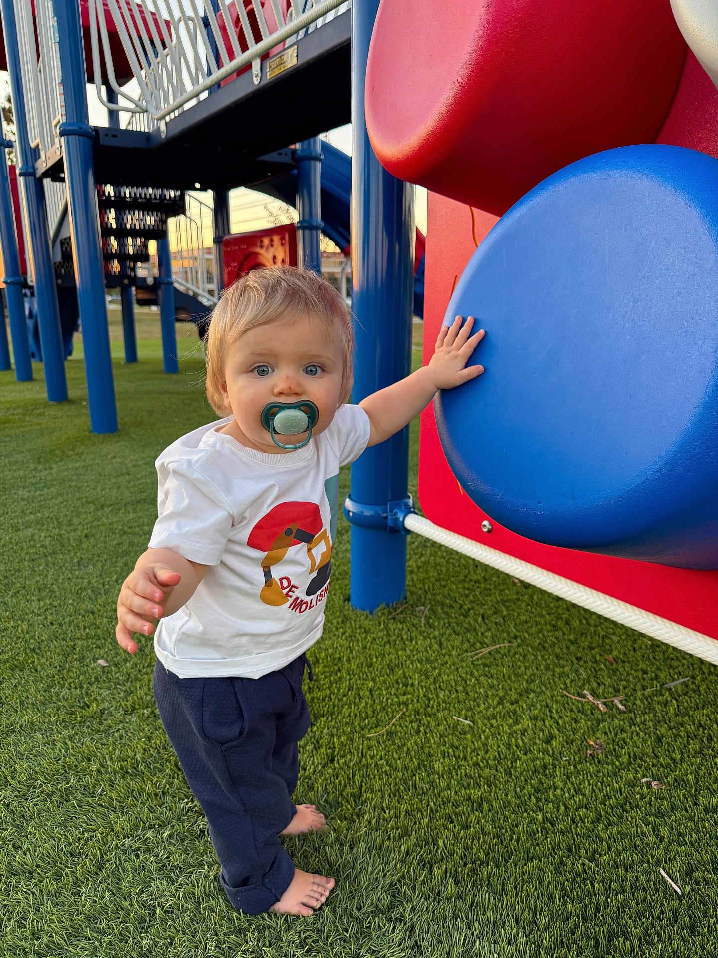 Kaiden is registered to the contest to win money with this photo: toddler, child, pacifier, barefoot, playground, play_equipment, grass, outdoor, blue, red, white_tshirt, pants, person, young_child, daylight, cute, curious, baby, exploring, fun