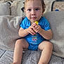 toddler, child, blue_onesie, couch, blanket, cushion, indoors, face, hands, feet, curious, sitting, patterned_fabric, soft_texture, young_child, portrait, baby, cozy, home, expression