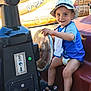 child, toddler, hat, blue_shirt, bib, smile, play_vehicle, steering_wheel, outdoor, sunlight, sandals, seat, happy, fun, summer, cute, young, play, toy, daytime