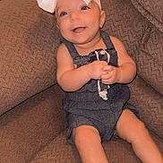Kelly is registered to the contest to win money with this photo: baby, child, smiling, bow, headwear, sitting, couch, furniture, indoors, happy, cute, infant, clothing, denim, barefoot, portrait, person, skin, expression, young