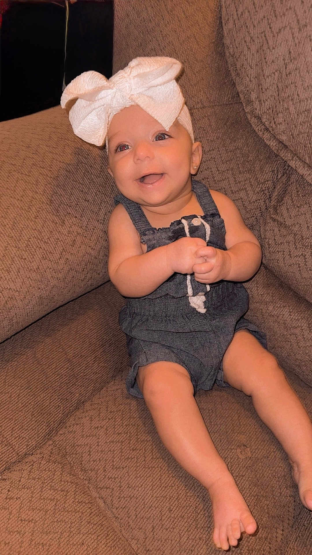 Kelly is registered to the contest to win money with this photo: baby, child, smiling, bow, headwear, sitting, couch, furniture, indoors, happy, cute, infant, clothing, denim, barefoot, portrait, person, skin, expression, young
