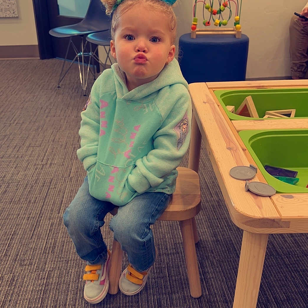 Trinity is registered to the contest to win money with this photo: baby, clothing, diningtable, face, footwear, furniture, hardwood, head, indoors, interiordesign, jeans, pants, person, photography, plywood, portrait, shoe, sitting, table, wood