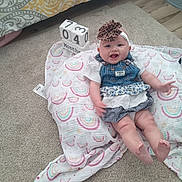 Raelyn is registered to the contest to win money with this photo: baby, infant, smiling, headband, bow, denim_dress, floral_pattern, checkered_pattern, blanket, rainbow_pattern, carpet, floor, block_calendar, months, cute, happy, child, indoors, portrait, clothing
