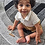 child, toddler, curly_hair, smiling, sitting, carpet, toy, playroom, indoors, white_shirt, shorts, barefoot, bracelet, floor, happy, cute, young_child, expression, person, home