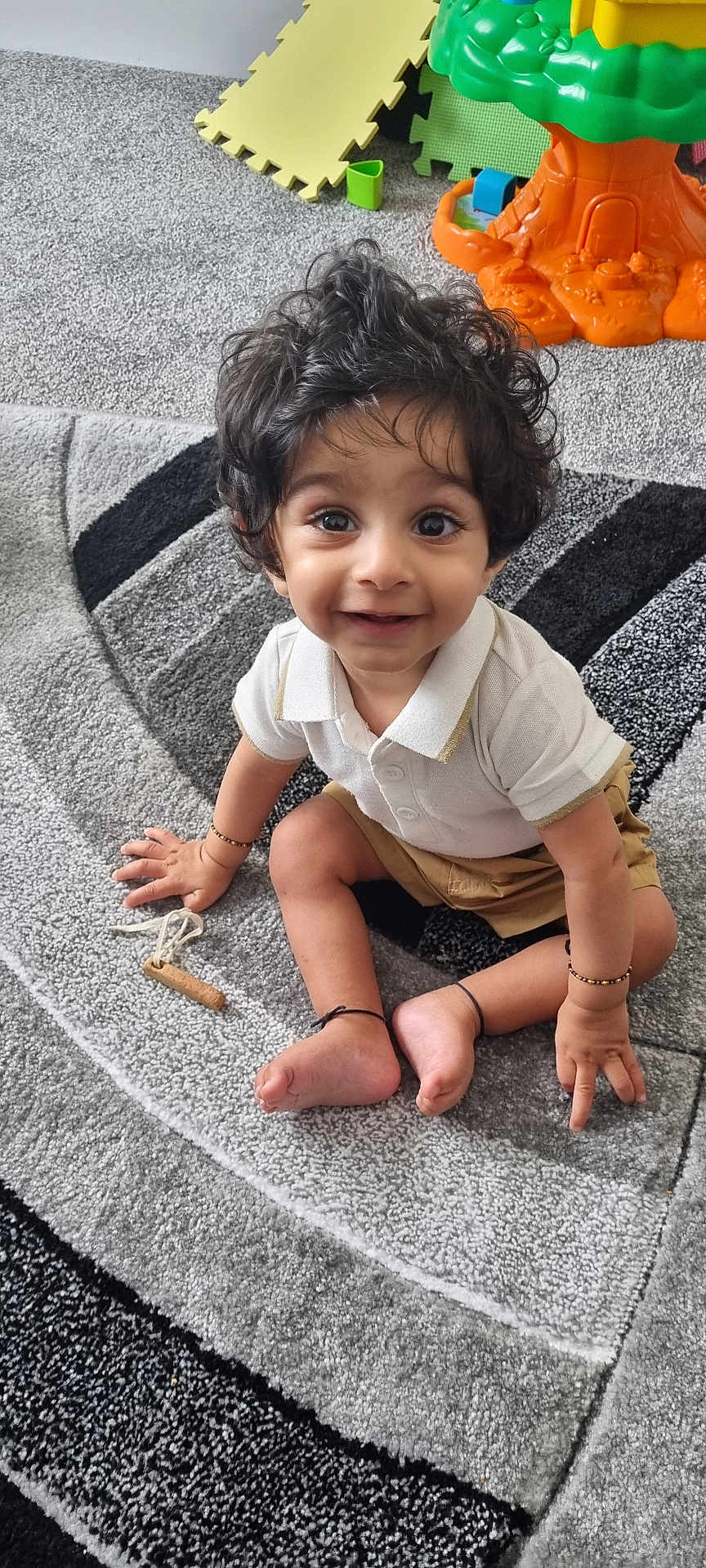 Viaan joined the competition — help win amazing prizes! child, toddler, curly_hair, smiling, sitting, carpet, toy, playroom, indoors, white_shirt, shorts, barefoot, bracelet, floor, happy, cute, young_child, expression, person, home