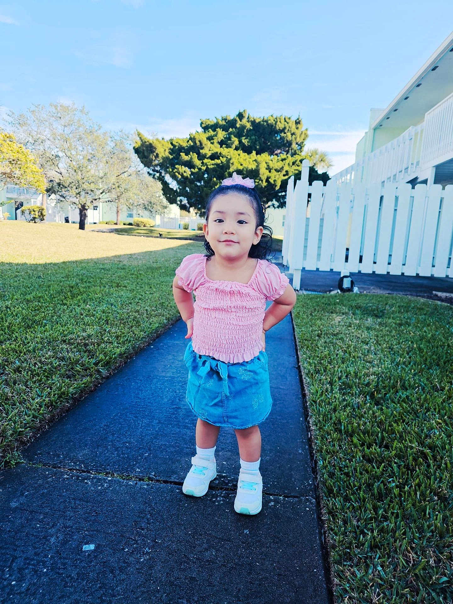 Adhara is registered to the contest to win money with this photo: child, girl, sidewalk, grass, pink_top, blue_skirt, white_sneakers, outdoor, daylight, smile, hair_bow, residential_area, trees, fence, concrete, playful, cute, young, portrait, standing