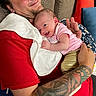 Abigail is registered to the contest to win money with this photo: baby, adult, tattoo, red_shirt, pink_shirt, gold_pants, couch, cushion, smile, happy, arm, hand, face, person, indoor, pattern, fabric, closeup, holding, cozy
