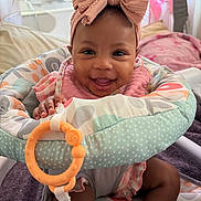 Ariah is registered to the contest to win money with this photo: baby, child, infant, smiling, happy, portrait, bow, headband, activity_seat, baby_toy, teething_ring, colorful, pink, indoor, bedroom, seated, face, eyes, clothing, playtime
