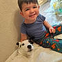 toddler, child, dog, smiling, sitting, blanket, indoor, flooring, wall, clothing, dinosaur_print, pet, happy, furniture, portrait, cute, cozy, young_child, animal, person
