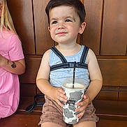 Theo joined the competition — help win amazing prizes! toddler, child, drink, cup, bench, wooden_bench, sitting, smiling, harness, shorts, tank_top, patterned_shorts, drink_cup, cow_print, happy, daylight, portrait, face, person, casual_clothing