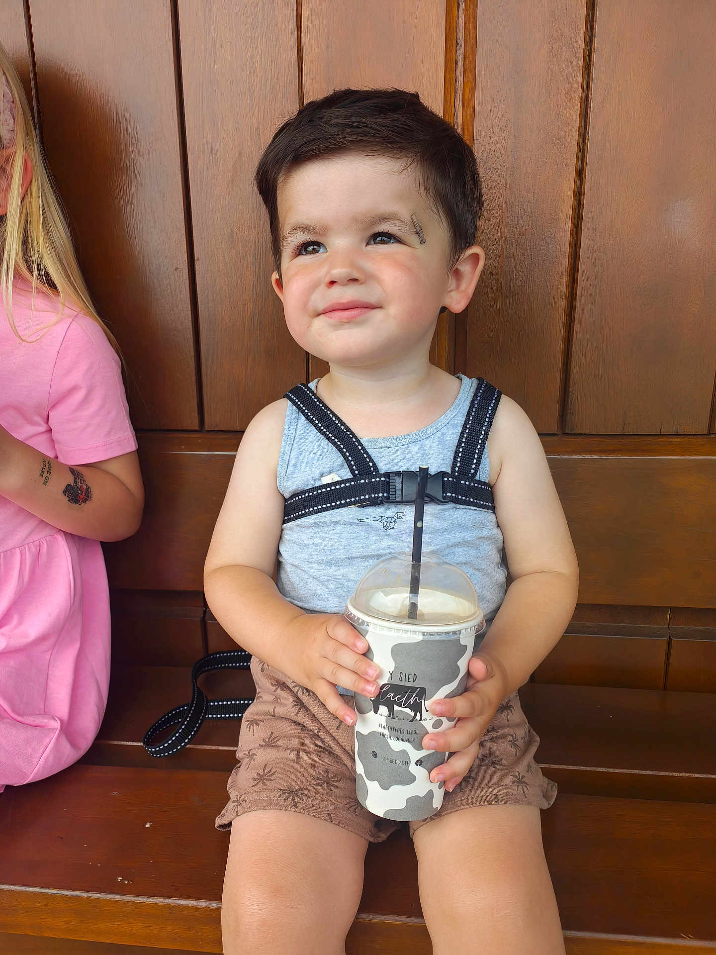 Theo joined the competition — help win amazing prizes! toddler, child, drink, cup, bench, wooden_bench, sitting, smiling, harness, shorts, tank_top, patterned_shorts, drink_cup, cow_print, happy, daylight, portrait, face, person, casual_clothing