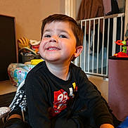 Theo is registered to the contest to win money with this photo: child, boy, smiling, indoor, toys, baby_gate, clothing, black_shirt, shoes, floor, coat, furniture, home, happy, portrait, casual, playtime, person, cute, young
