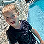 child, boy, smiling, pool, water, swimwear, dinosaur, shorts, black_shirt, stone_wall, sunlight, outdoor, summer, happy, wet_hair, blue_sky, trees, playful, leisure, vacation