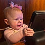 baby, child, pink_bow, hands, wooden_table, black_device, curious, indoor, wood_paneling, window, natural_light, face, portrait, sitting, person, infant, cute, expression, close_up, baby_girl
