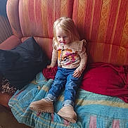 Lina participe au concours pour gagner de l'argent avec cette photo : blanket, boots, casual_clothing, child, couch, floor, indoor, jeans, light_blond_hair, long_hair, pacifier, pillow, portrait, red_blanket, relaxed, sitting, striped_couch, toy_design, wall, young_child