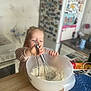 toddler, child, kitchen, mixing_bowl, whisk, flour, countertop, appliance, refrigerator, magnet, stove, cooking, baking, utensil, person, indoor, clothing, table, food_preparation, home