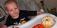 Teddy joined the competition — help win amazing prizes! baby, child, high_chair, pumpkin, orange, white, yellow, smile, face, clothing, black_shirt, indoor, furniture, soft_cover, table, toy, person, cute, festive, fall