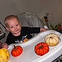 baby, child, high_chair, pumpkin, orange, white, yellow, smile, face, clothing, black_shirt, indoor, furniture, soft_cover, table, toy, person, cute, festive, fall