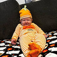 Teddy is registered to the contest to win money with this photo: baby, infant, orange_hat, striped_pajamas, pumpkin, smiling, couch, blanket, ghost_pattern, halloween_theme, footed_pajamas, cute, indoor, child, festive, cozy, orange, happy, portrait, holiday